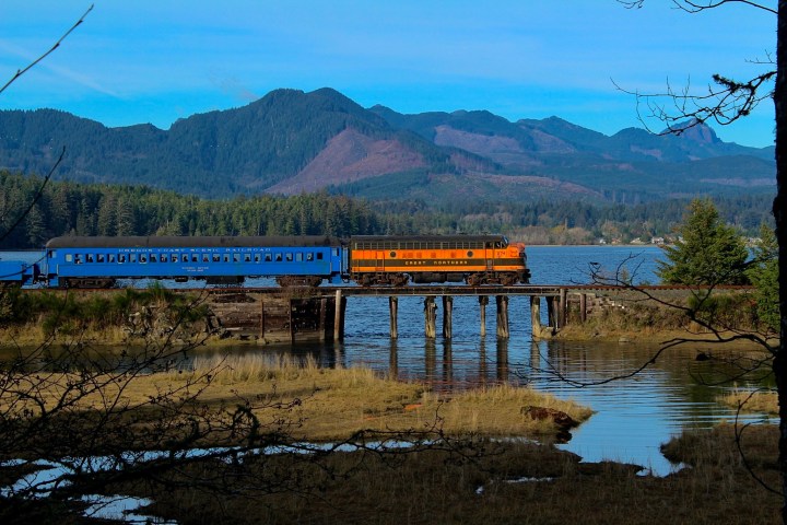 Train crossing a bridge over water with mountains in the background and trees in the foreground.