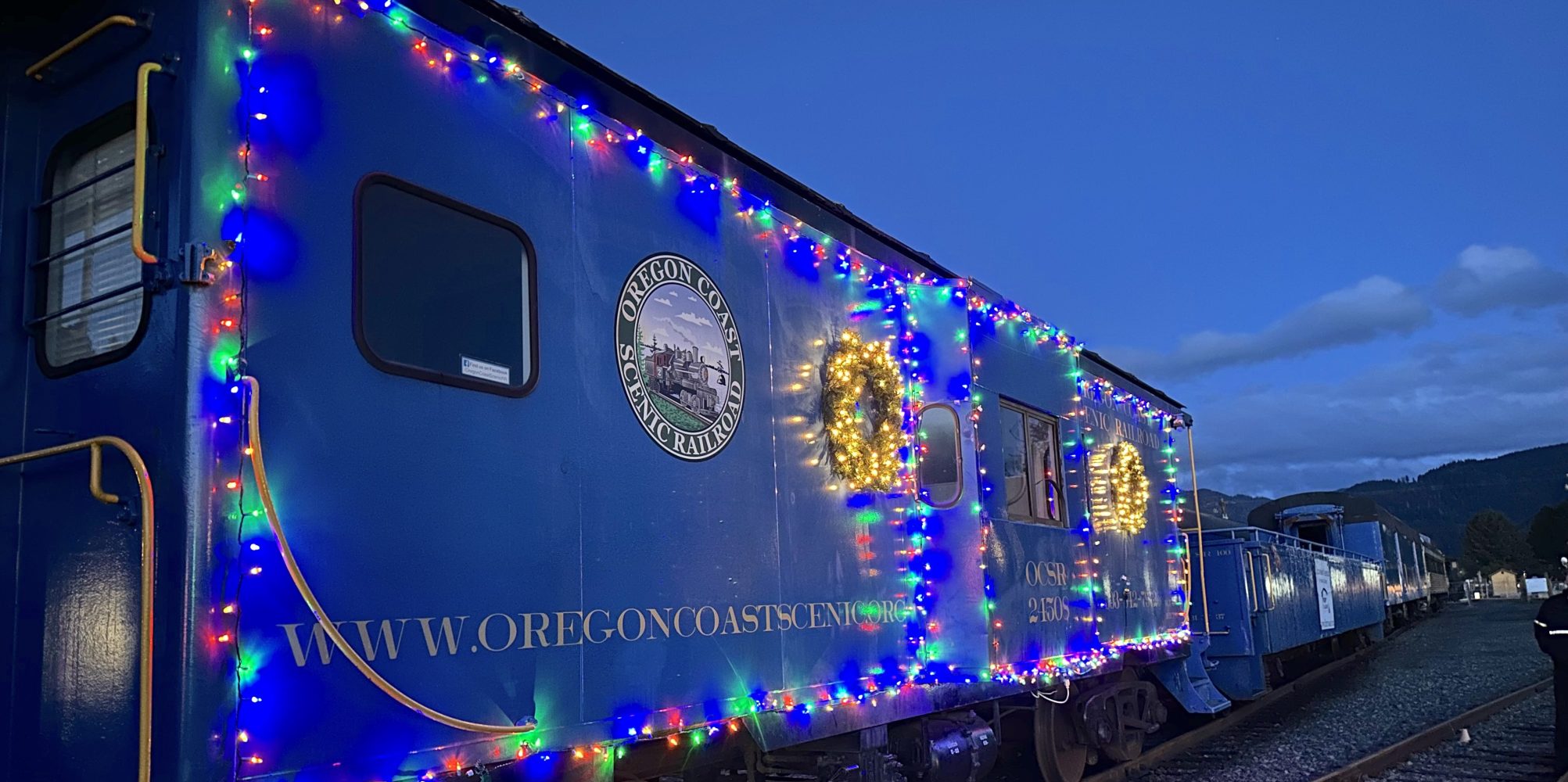 Blue train car decorated with colorful lights and wreaths at dusk on railway.