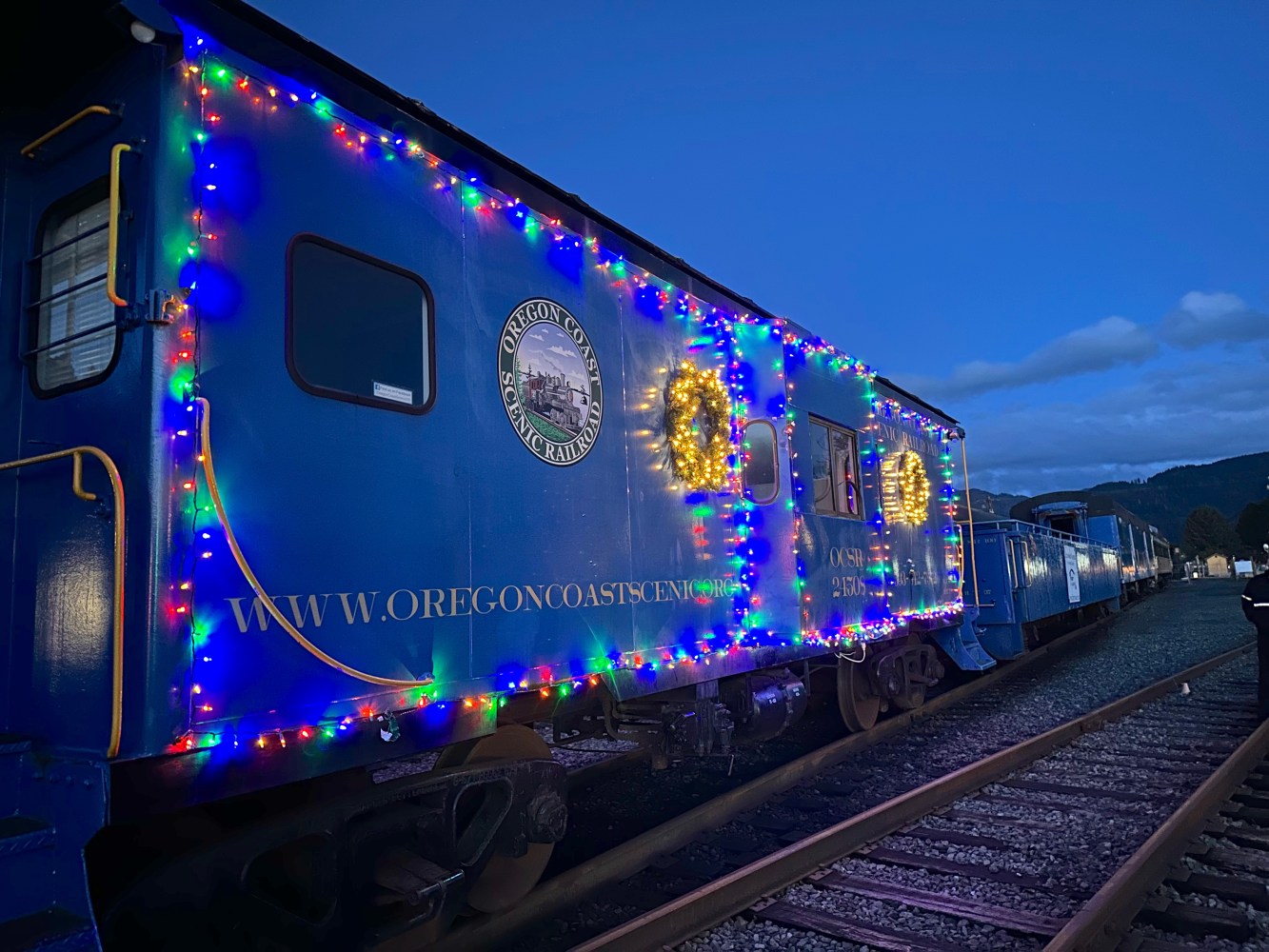 Blue train car decorated with colorful lights and wreaths at dusk on railway.