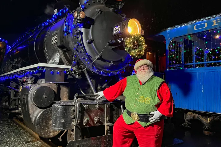 Santa Claus standing by a decorated steam locomotive at night, with festive lights and a wreath.