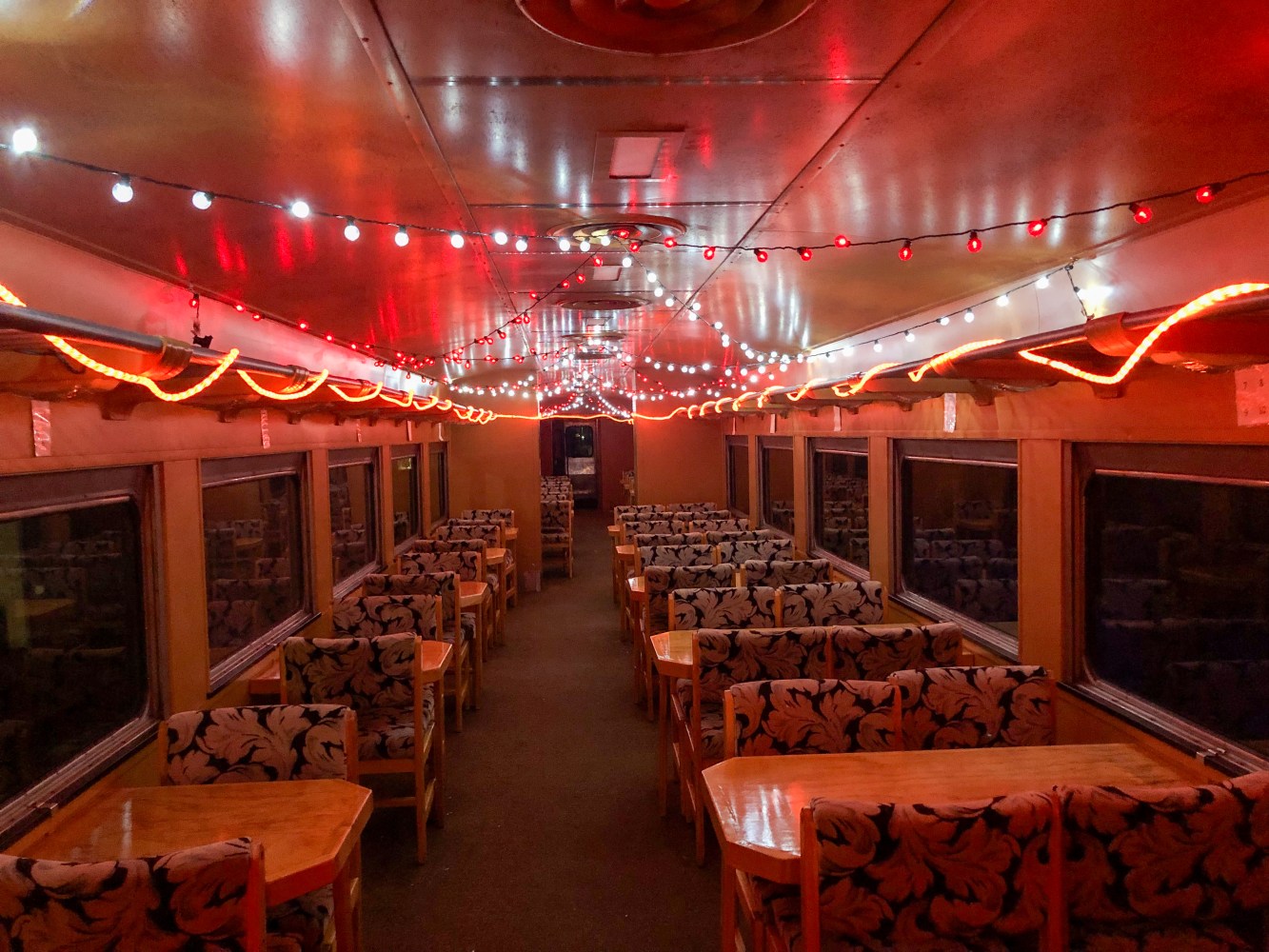 Train dining car with red and white string lights, floral chairs, and empty wooden tables.