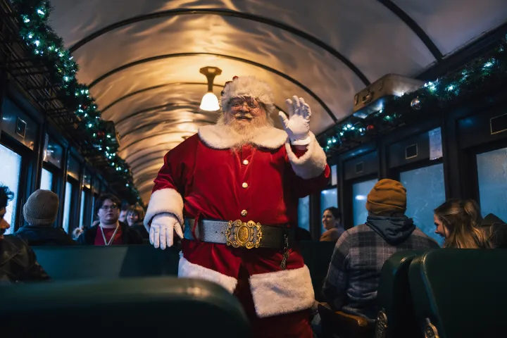 Santa waving inside a festively decorated train with passengers seated around.