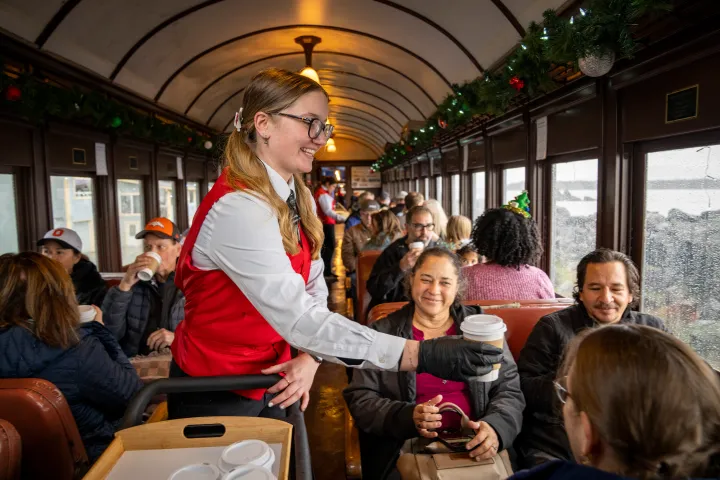 Train staff serving drinks to seated passengers inside a decorated train car.