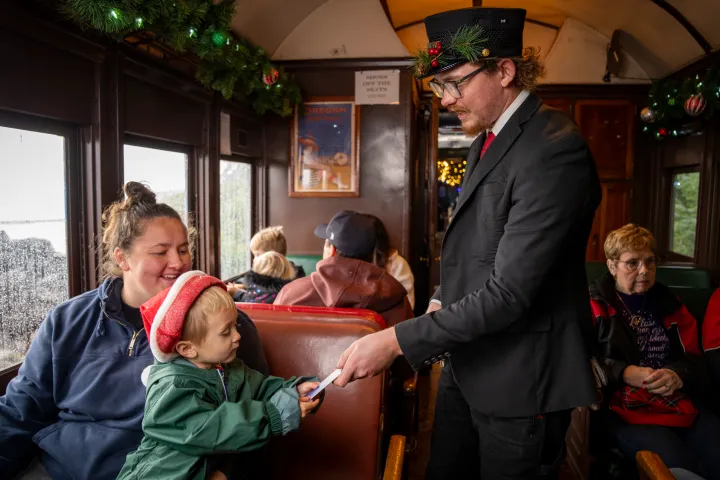 Conductor in holiday attire checks ticket of child in festive hat on train.