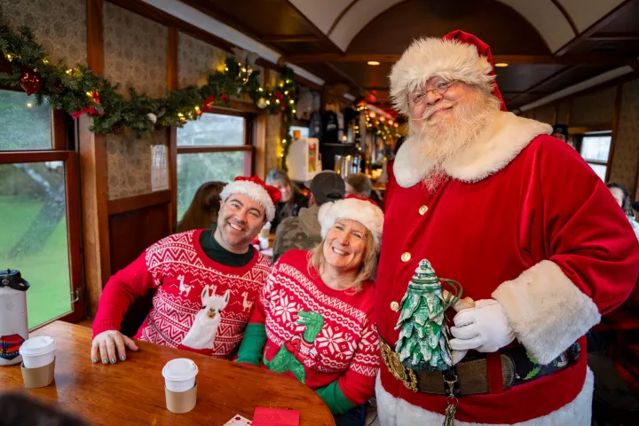 Santa with two people in Christmas sweaters on a train with decorations and coffee cups.