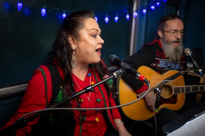 Two musicians performing, woman singing, man playing guitar, festive sweaters, blue lights in background.
