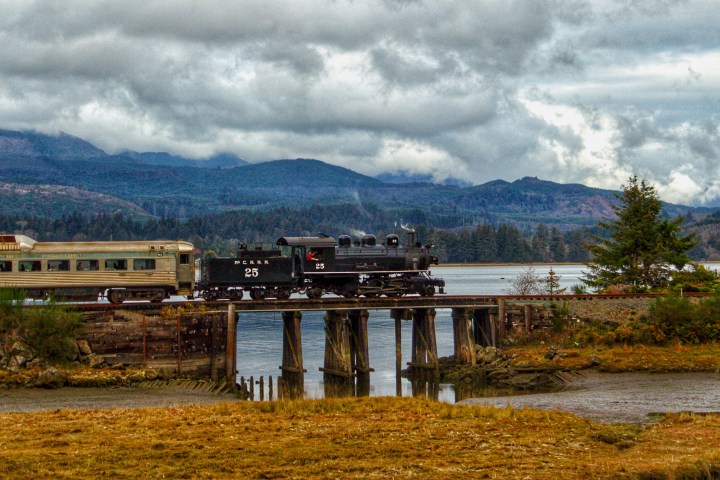 Steam train crossing a wooden bridge over a river with mountains in the background.