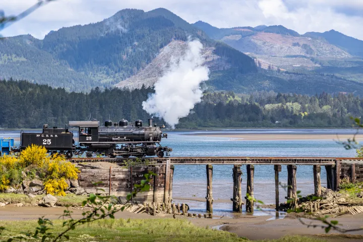 Steam train crossing a wooden bridge over water with mountains in the background.