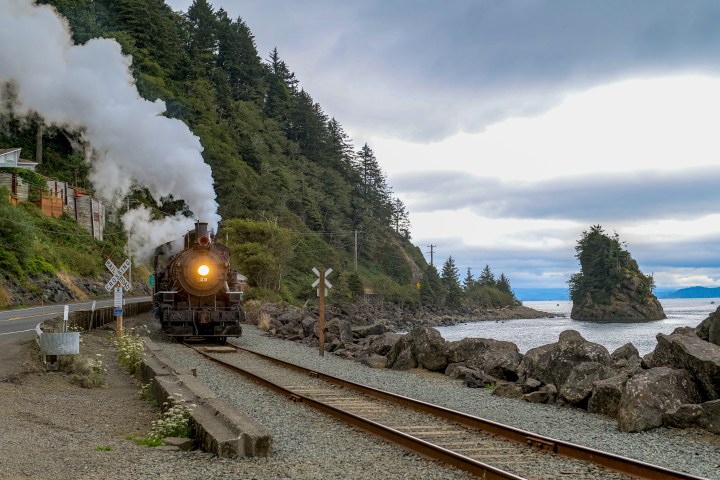 Steam train on coastal track with rocky shoreline and trees.