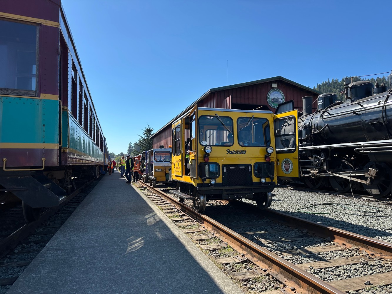 Train station with parked trains and yellow maintenance vehicles on tracks under clear blue sky.
